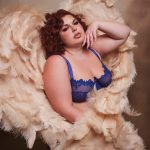 A woman in blush feather wings and blue lingerie poses in a fantasy boudoir session against a soft cream backdrop.